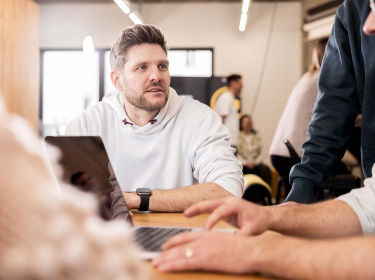 Mehrere Personen sitzen an einem Tisch und arbeiten gemeinsam an Laptops, dabei findet ein Gespräch oder eine Diskussion statt.
