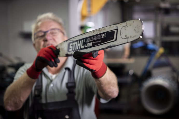 Motorsägen Wartung Ein Mann mit Schutzbrille und roten Handschuhen hält eine Kettensäge mit der Aufschrift „STIHL“ in die Kamera, wobei der Fokus auf der Säge liegt.