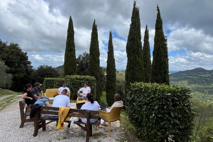 AB3 Team Workation in der Toskana Das AB3-Team sitzt auf Gartenmöbeln unter hohen Zypressen und arbeitet im Freien, mit einer grünen Hügel-Landschaft im Hintergrund.