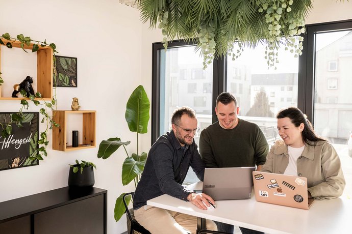 Drei Personen sitzen an einem Tisch in einem hellen Büro und schauen gemeinsam auf zwei offene Laptops. Sie wirken konzentriert und arbeiten zusammen an einem Projekt. Im Hintergrund sind Pflanzen und moderne Dekoration sichtbar.