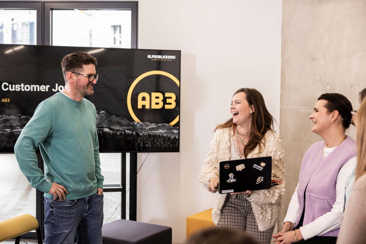 Drei Personen unterhalten sich in einem modernen Büroraum freundlich und lächelnd vor einem Bildschirm mit der Aufschrift „Customer Journey AB3“ in der Werbeagentur in Tettnang.
