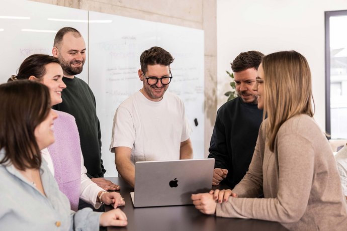 Sechs Menschen stehen in einem hellen Büro um einen Laptop und besprechen konzentriert ein gemeinsames Projekt in der Werbeagentur in Tettnang.