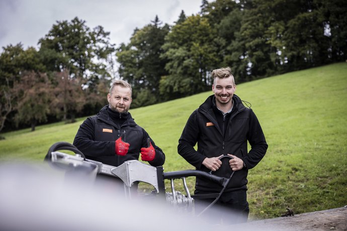 Zwei Mitarbeiter von Bentele bei der Arbeit Zwei Männer in schwarzer Arbeitskleidung stehen auf einer Wiese, lächeln und geben Daumen hoch. Vor ihnen befindet sich Arbeitsgerät. Im Hintergrund Bäume und ein bewölkter Himmel.