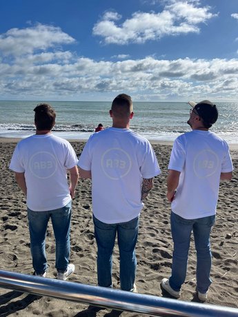 AB3 Team in Teamshirts am Strand Drei Mitglieder des Alpenblickdrei-Teams blicken auf das Meer, mit weißen T-Shirts und dem AB3-Logo, vor einem klaren blauen Himmel und der Küste.