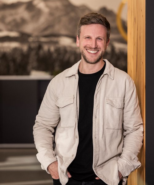 Simon Bauer Alpenblickdrei Porträt eines lächelnden Mannes mit leichter Bartstoppel, der entspannt an eine Wand gelehnt steht, trägt eine helle Jacke über schwarzem Shirt.