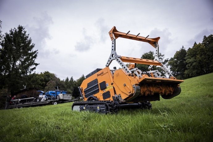 Landtechnik von Bentele in Baindt Ein orangefarbener, ferngesteuerter Mähroboter steht auf einer grasbewachsenen Wiese, im Hintergrund sind Bäume und ein blauer Traktor zu sehen.