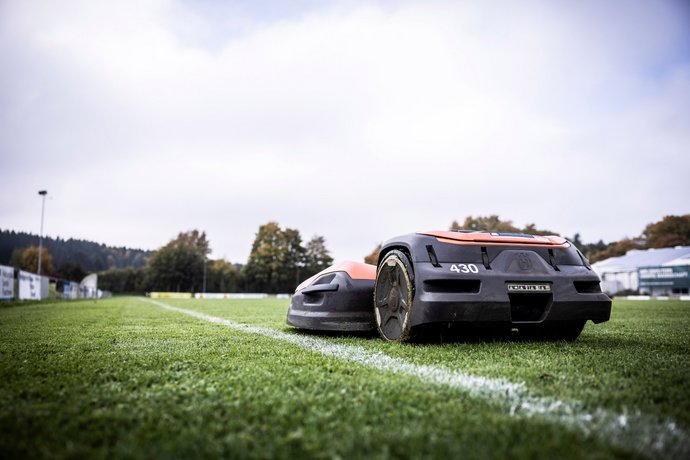 Mähroboter für Sportplatz Zwei autonome Rasenmähroboter fahren auf einem gepflegten Fußballfeld nebeneinander entlang einer weißen Spielfeldlinie.
