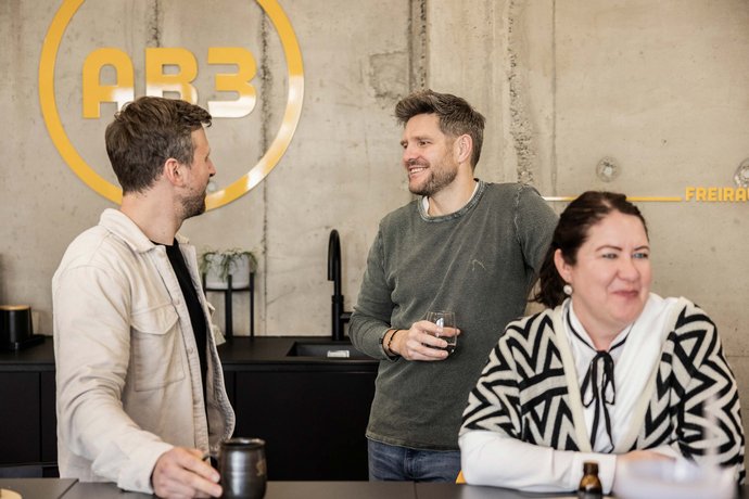Drei Personen unterhalten sich locker in einem modernen Büro mit Betonwänden, zwei Männer stehen, eine Frau sitzt am Tresen.