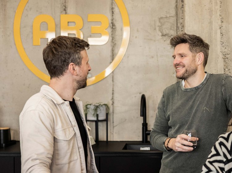 Drei Personen unterhalten sich locker in einem modernen Büro mit Betonwänden, zwei Männer stehen, eine Frau sitzt am Tresen.