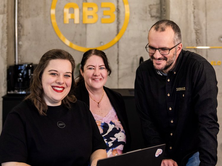 Casual Teamfoto im Industrie-Stil Drei Mitarbeitende der Alpenblickdrei Werbeagentur sitzen entspannt vor Betonwand in kreativem Arbeitsumfeld.