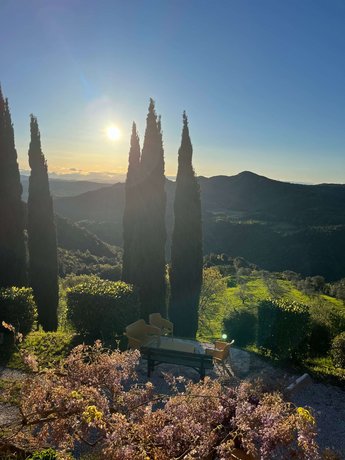 Sonnenuntergang in der Toskana Ausblick aus unserer Unterkunft I Casali Toscana, Blick auf einen idyllischen Garten mit Zypressen und Sitzgelegenheiten, vor einer hügeligen Landschaft im Sonnenuntergang.