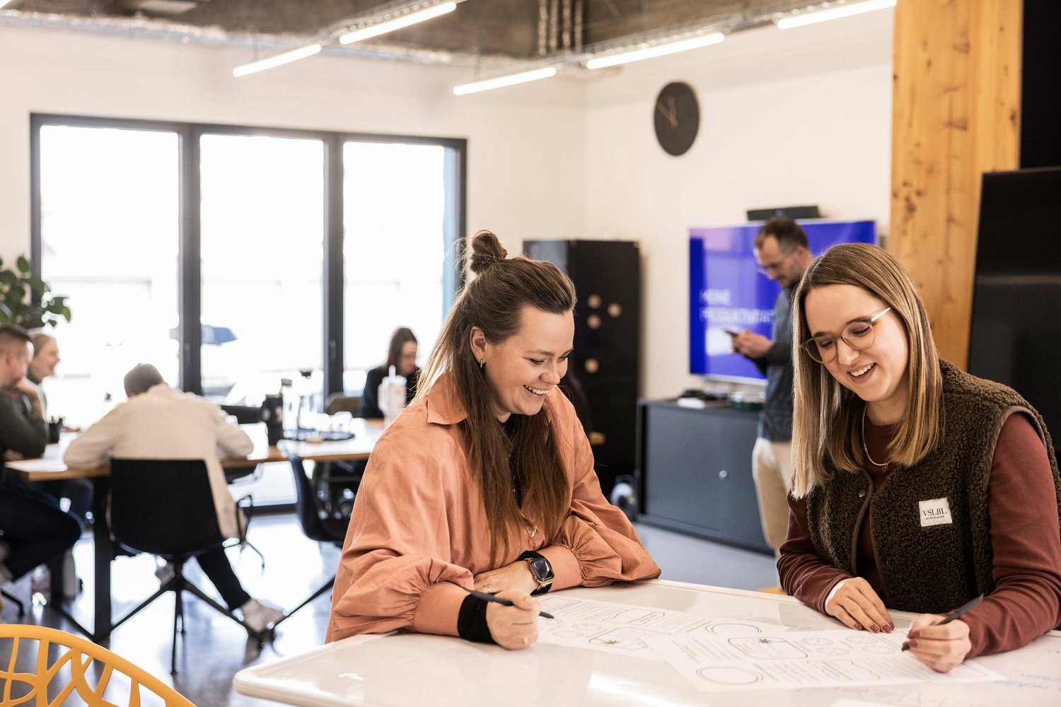 Zwei Frauen arbeiten gemeinsam an einem Projektplan oder einer Zeichnung an einem Tisch in einem modernen, hellen Büro. Im Hintergrund sind weitere Personen in konzentrierter Arbeitsatmosphäre zu sehen.