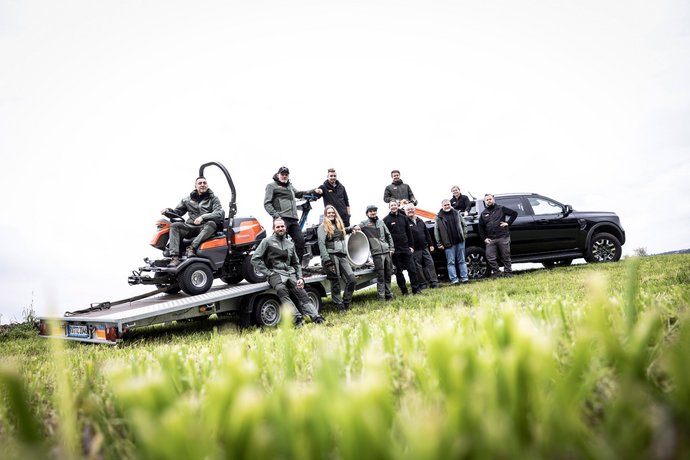 Team von Bentele Forst- und Gartentechnik Eine Gruppe von elf Menschen steht und sitzt auf einem Anhänger, auf dem ein Rasenmäher sowie weitere Ausrüstungsgegenstände geladen sind, neben einem schwarzen Auto auf einer Wiese. Die Szene wirkt professionell und gemeinschaftlich.