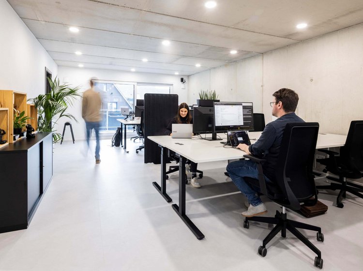 Moderner, heller Büroraum mit mehreren Personen, die an Tischen an Computern arbeiten. Marvin Lang sitzt an einem Schreibtisch, Katharina Reitemann ist mit Laptop beschäftigt, eine weitere Person geht im Hintergrund vorbei.