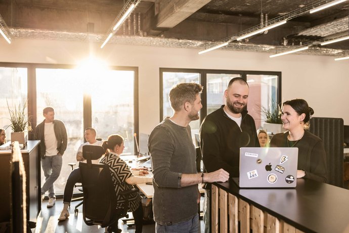 Drei Kollegen stehen im Büro in der Werbeagentur ALPENBLICKDREI in Tettnang an einem Stehpult, lachen und schauen auf einen Laptop.
