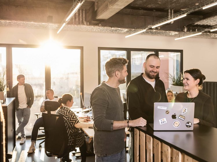 Drei Kollegen stehen im Büro in der Werbeagentur ALPENBLICKDREI in Tettnang an einem Stehpult, lachen und schauen auf einen Laptop.