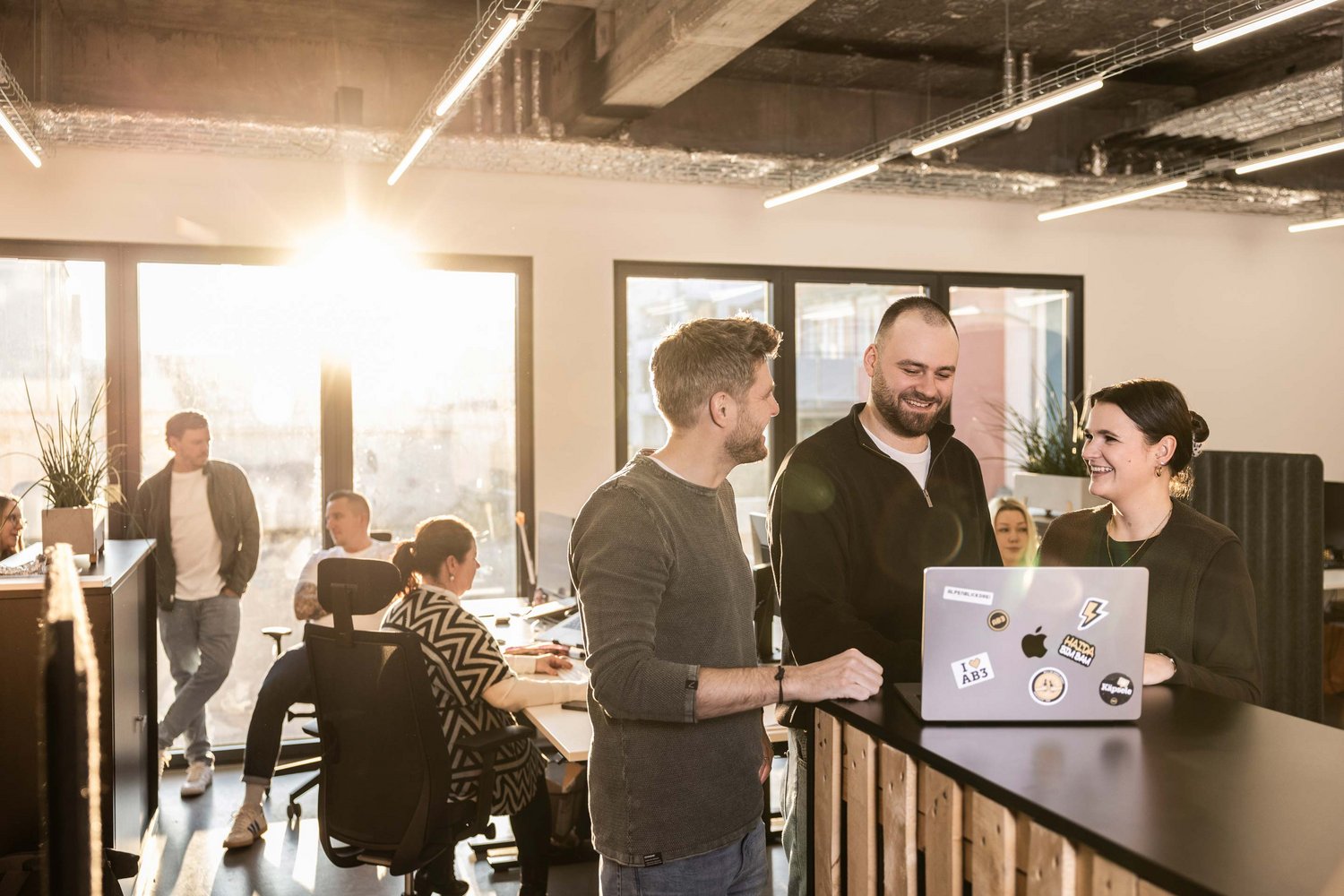 Drei Kollegen stehen im Büro in der Werbeagentur ALPENBLICKDREI in Tettnang an einem Stehpult, lachen und schauen auf einen Laptop.