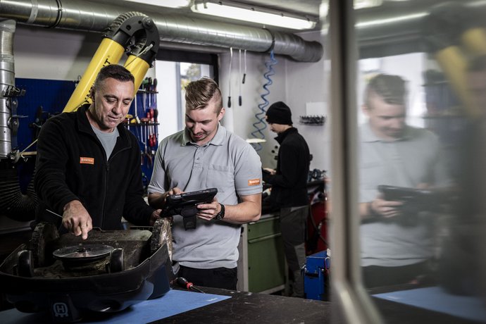 Zwei Mitarbeiter von Bentele bei der Arbeit Zwei Männer arbeiten gemeinsam in einer Werkstatt, einer prüft ein Teil, der andere hält ein Tablet, um Informationen zu überprüfen. Im Hintergrund arbeitet eine dritte Person an einer anderen Aufgabe.