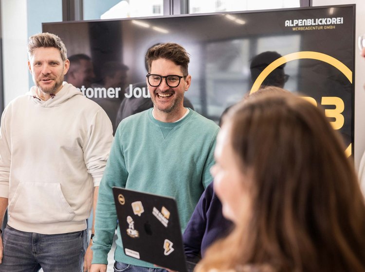 Mehrere Personen unterhalten sich locker und lächelnd in einem modernen Büro vor einem Bildschirm mit dem Logo der Werbeagentur Alpenblickdrei.