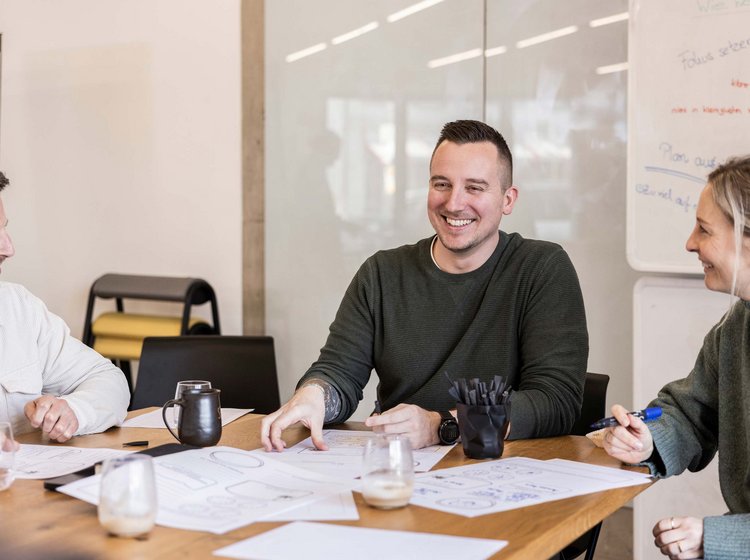 Drei Personen sitzen an einem Tisch, diskutieren und lächeln während eines Meetings. Auf dem Tisch liegen Papiere mit Skizzen, und im Hintergrund ist ein Whiteboard mit handschriftlichen Notizen zu sehen.