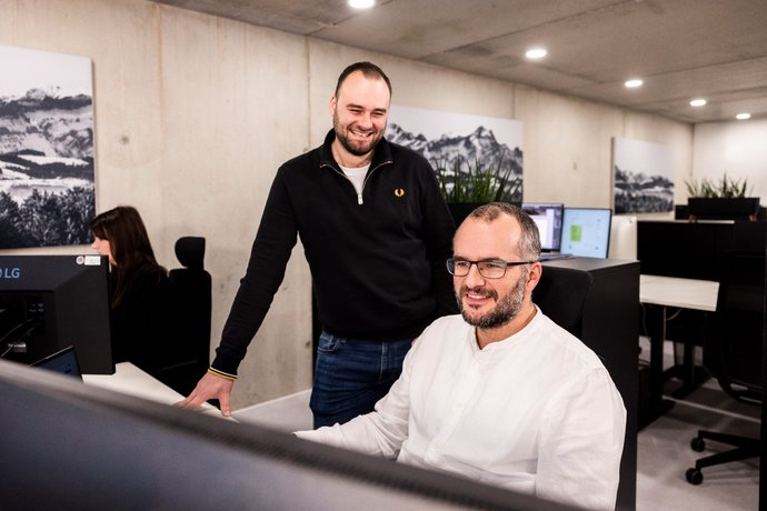 Kollegen im Dialog Zwei Männer der Alpenblickdrei Werbeagentur im Gespräch am Arbeitsplatz, einer steht, der andere sitzt.