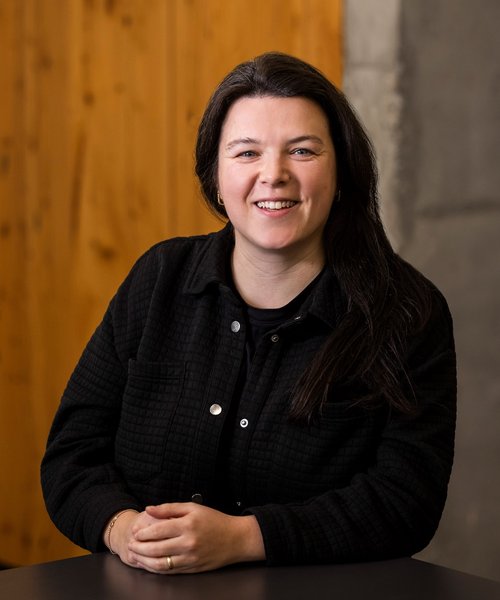 Selina Ruepp Alpenblickdrei Porträt einer lächelnden Frau mit langen dunklen Haaren, die an einem Tisch sitzt und in die Kamera schaut. Sie trägt eine schwarze Jacke und eine entspannte Haltung.