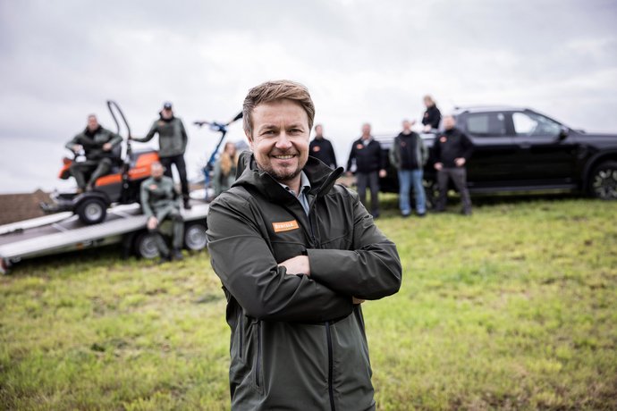 Geschäftsführer von Bentele Forst- und Gartentechnik Ein lächelnder Mann steht mit verschränkten Armen auf einer Wiese, im Hintergrund mehrere Personen, Fahrzeuge und ein Anhänger mit Arbeitsgeräten. Alle tragen Outdoor-Kleidung, was auf ein Team in einem Arbeits- oder Außeneinsatz hinweist.