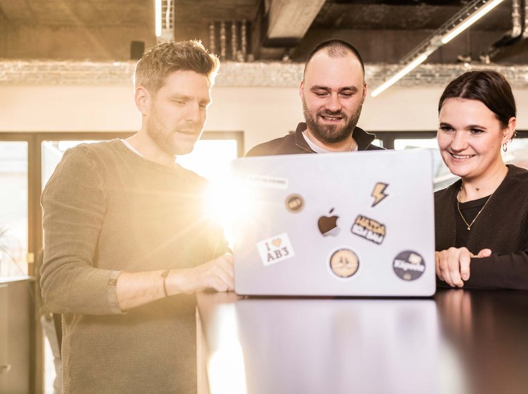 Drei Personen arbeiten gemeinsam an einem Laptop und lächeln dabei, in einem modernen, hellen Büro.