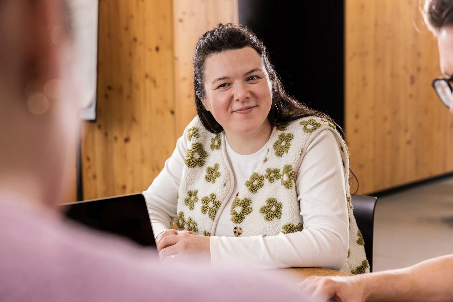 Eine lächelnde Frau sitzt an einem Tisch und blickt aufmerksam auf Gesprächspartner, während sie an einer Besprechung teilnimmt.