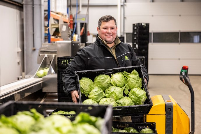 Landwirt mit Salatköpfen Ein junger Landwirt hält eine Kiste mit Salatköpfen, im Hintergrund landwirtschaftliche Geräte in einer Halle.